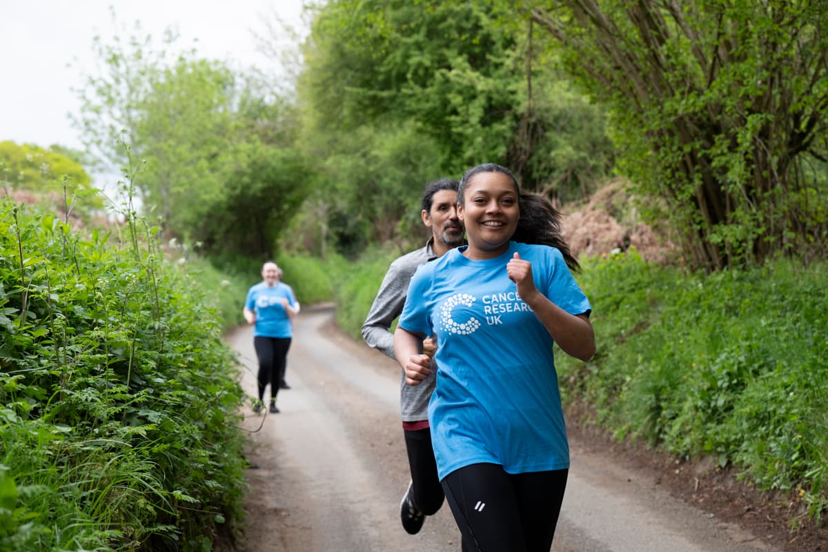 Three people running on a rural path in blue Cancer Research UK shirts, surrounded by green foliage.