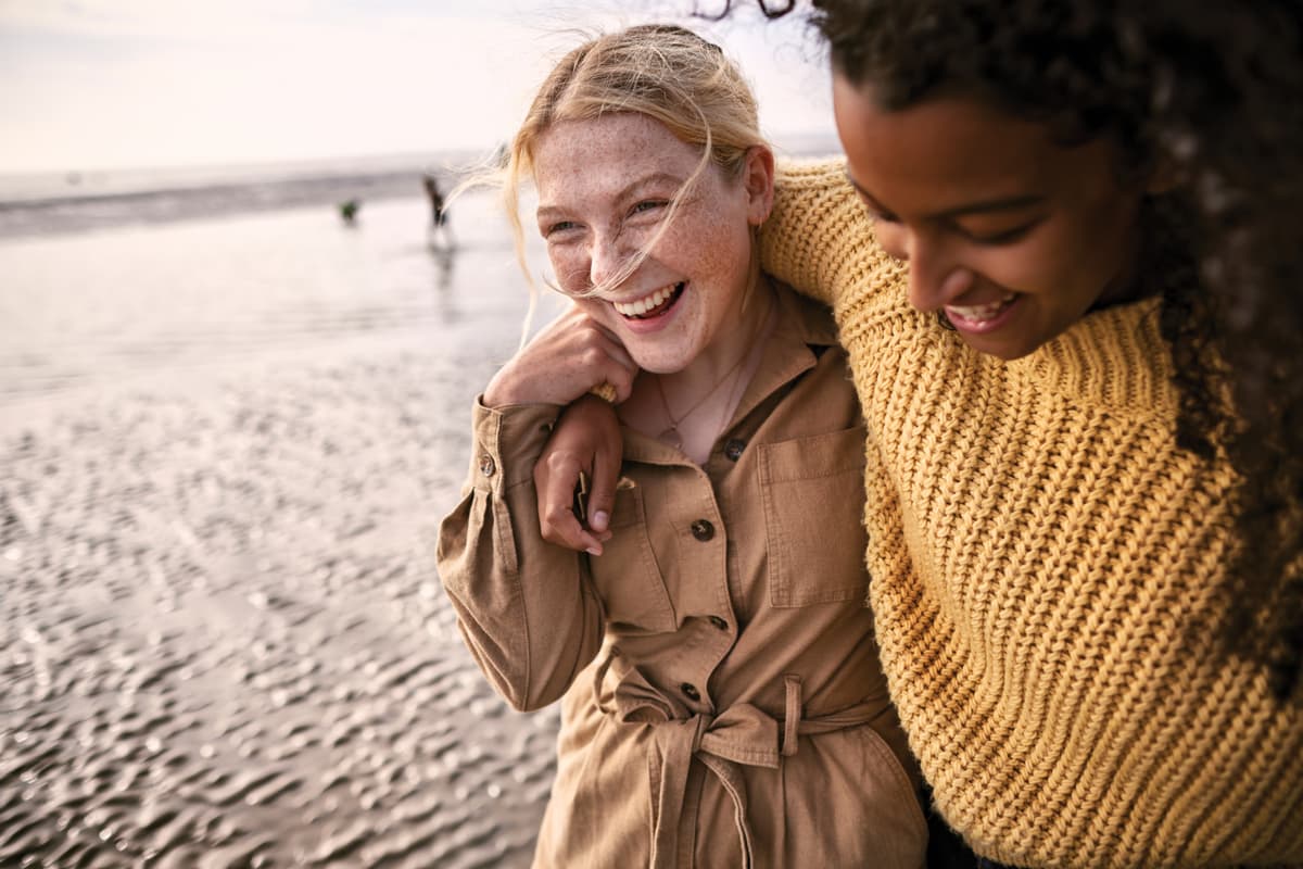 A photo of two women walking on the beach laughing and hugging.