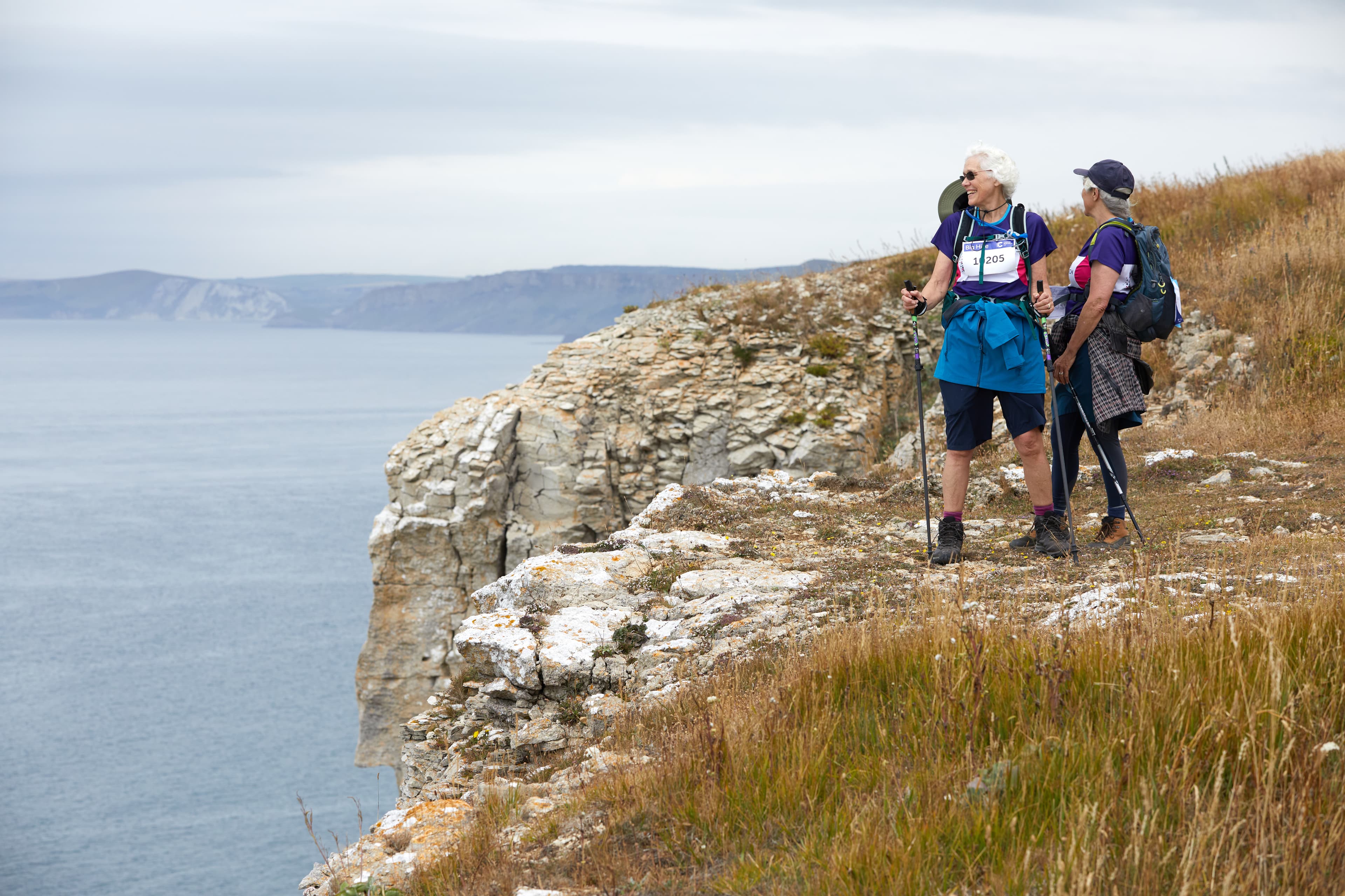Two Big Hike participants stand on a rocky cliff overlooking the sea, both wearing CRUK shirts and using walking poles.