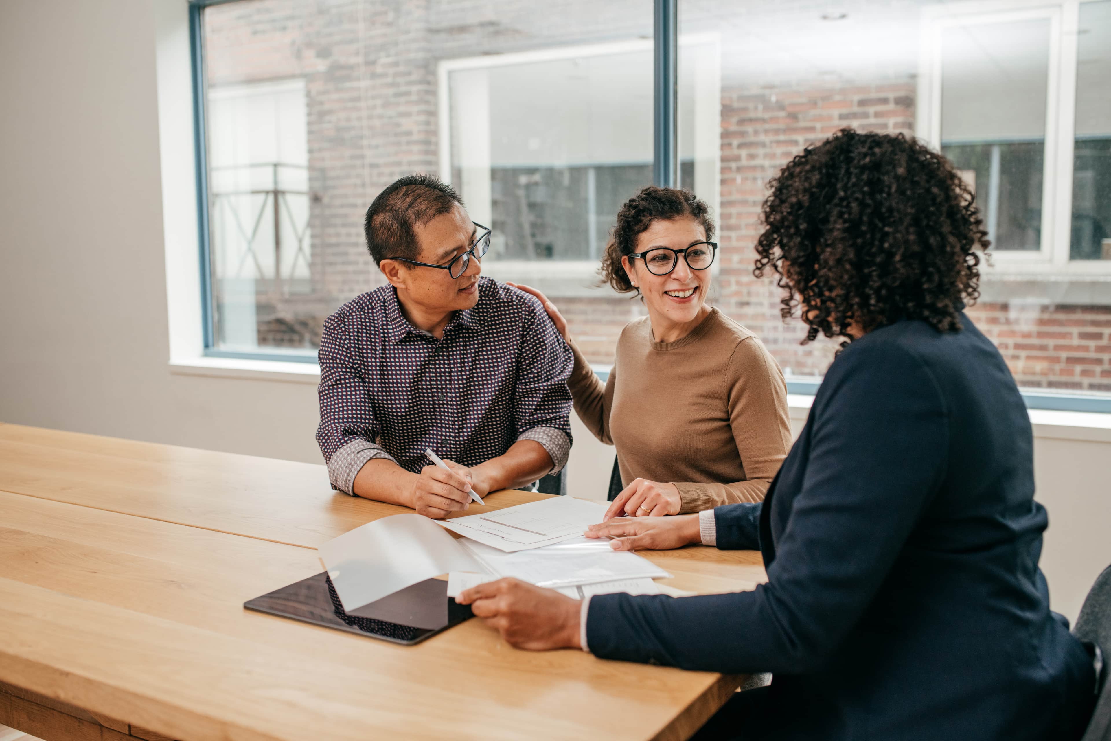 A couple smiling as they sign a document. They are sat at a table with a legal professional.