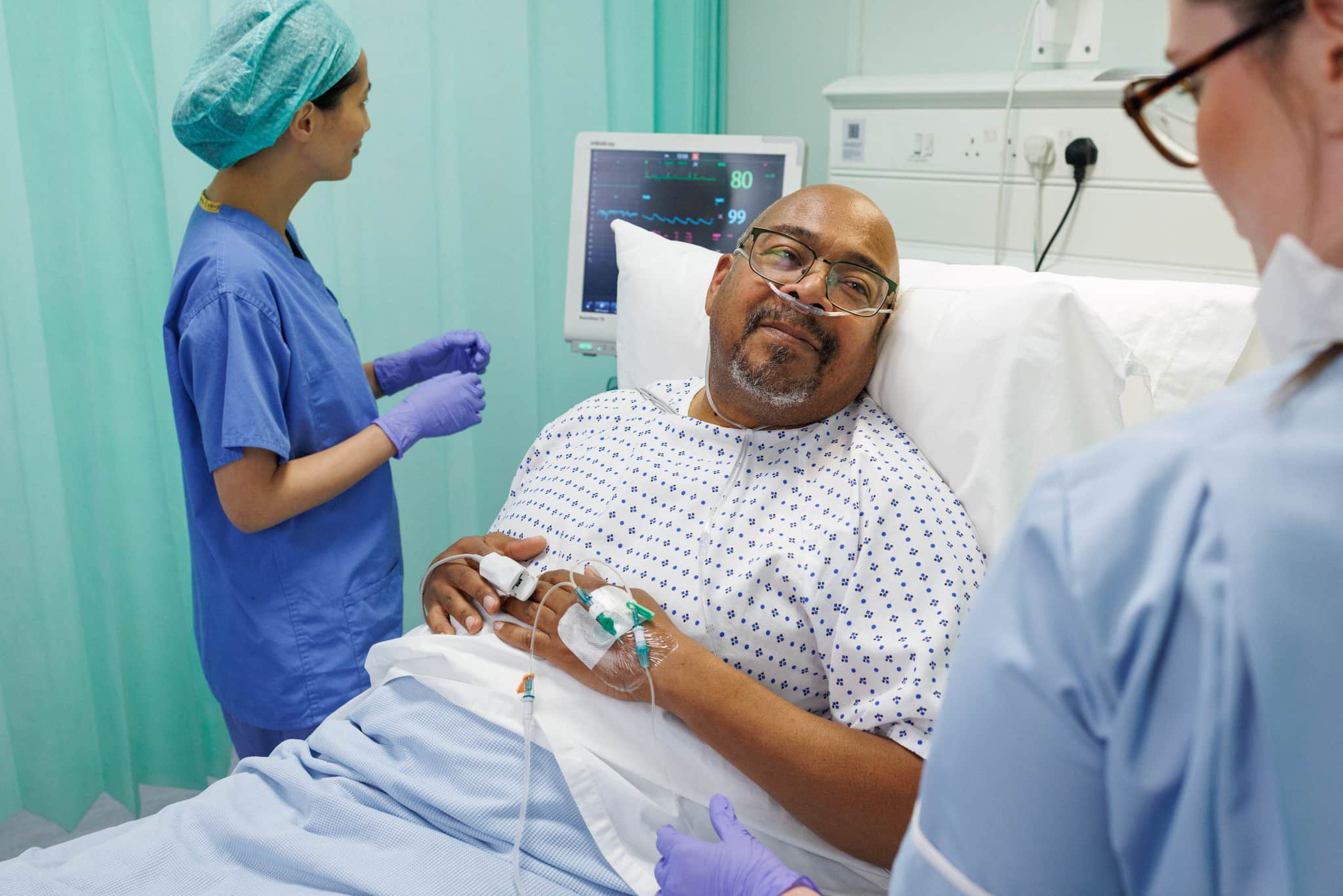 Patient on hospital bed interacting with a nurse while a doctor reads from a bedside monitor.