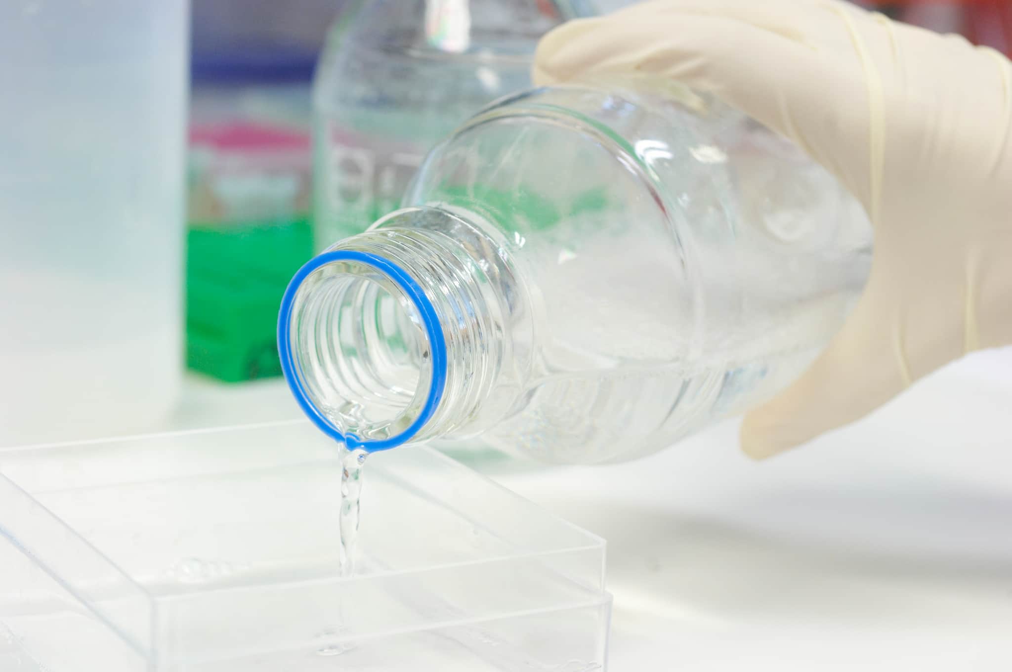 Clear liquid being poured from a Duran bottle into a square container in a lab.