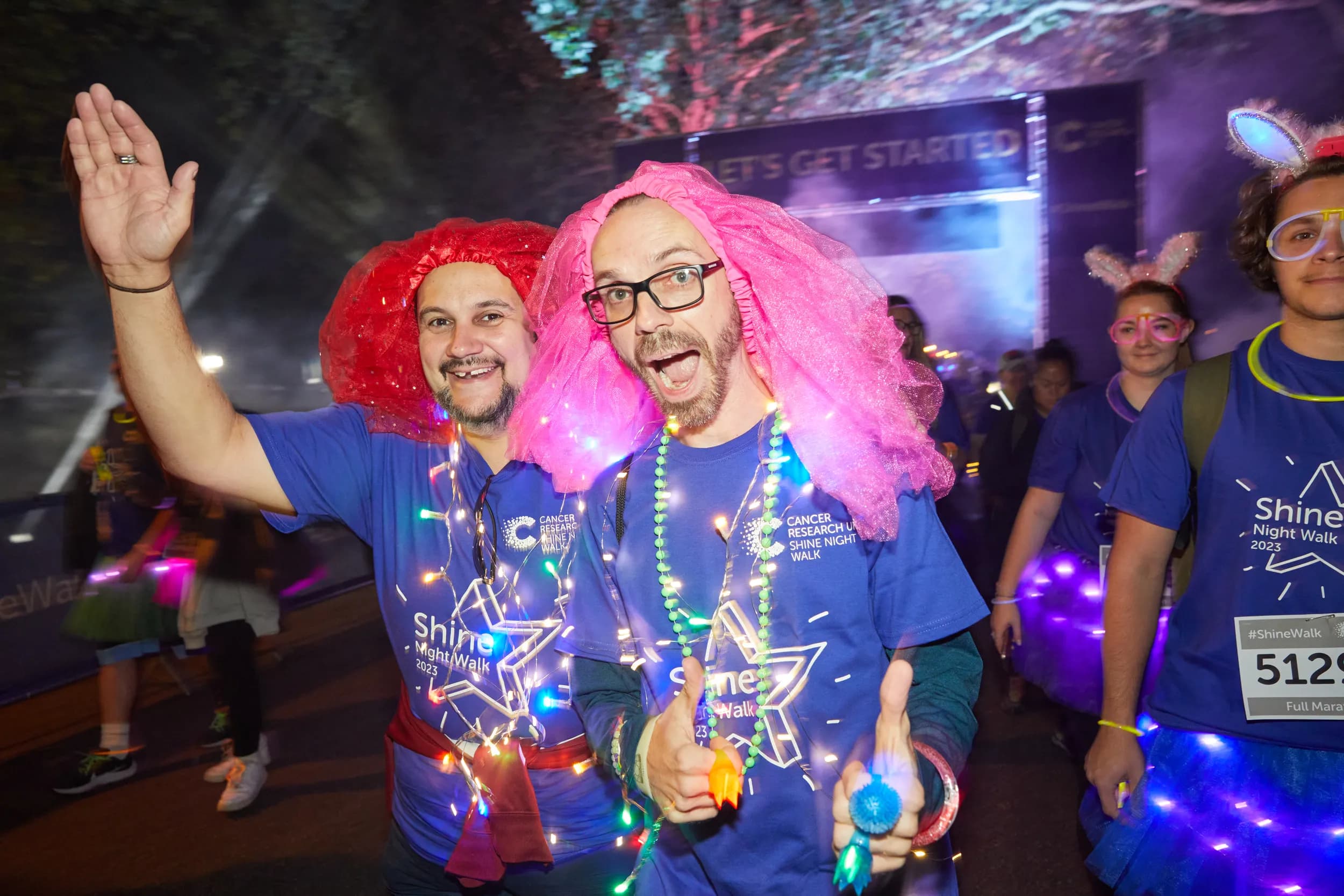 Two men wearing colourful accessories waving to the camera as they walk through the start point at Shine Night Walk.
