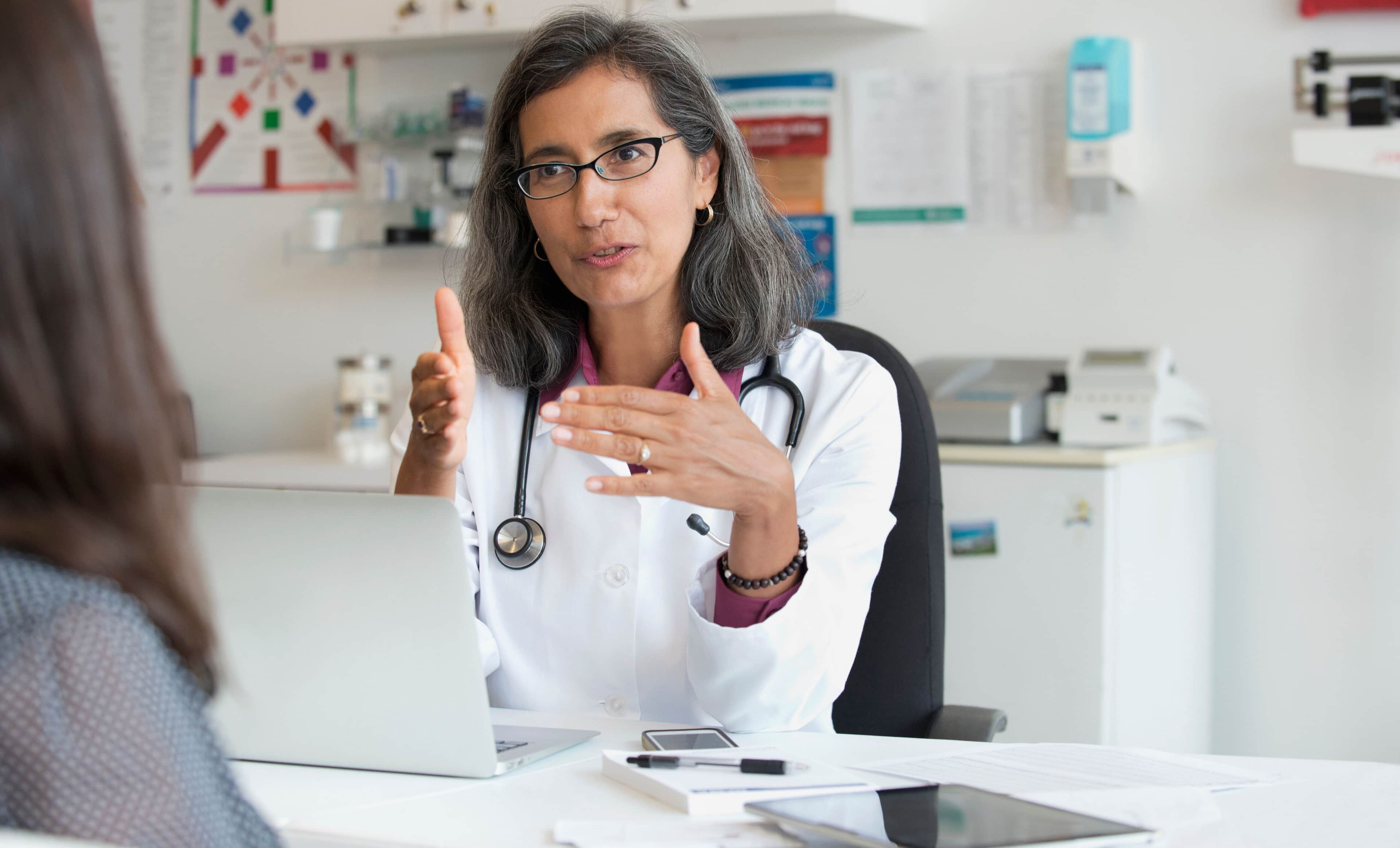 Doctor wearing a stethoscope with a laptop in front of them is talking to a patient.