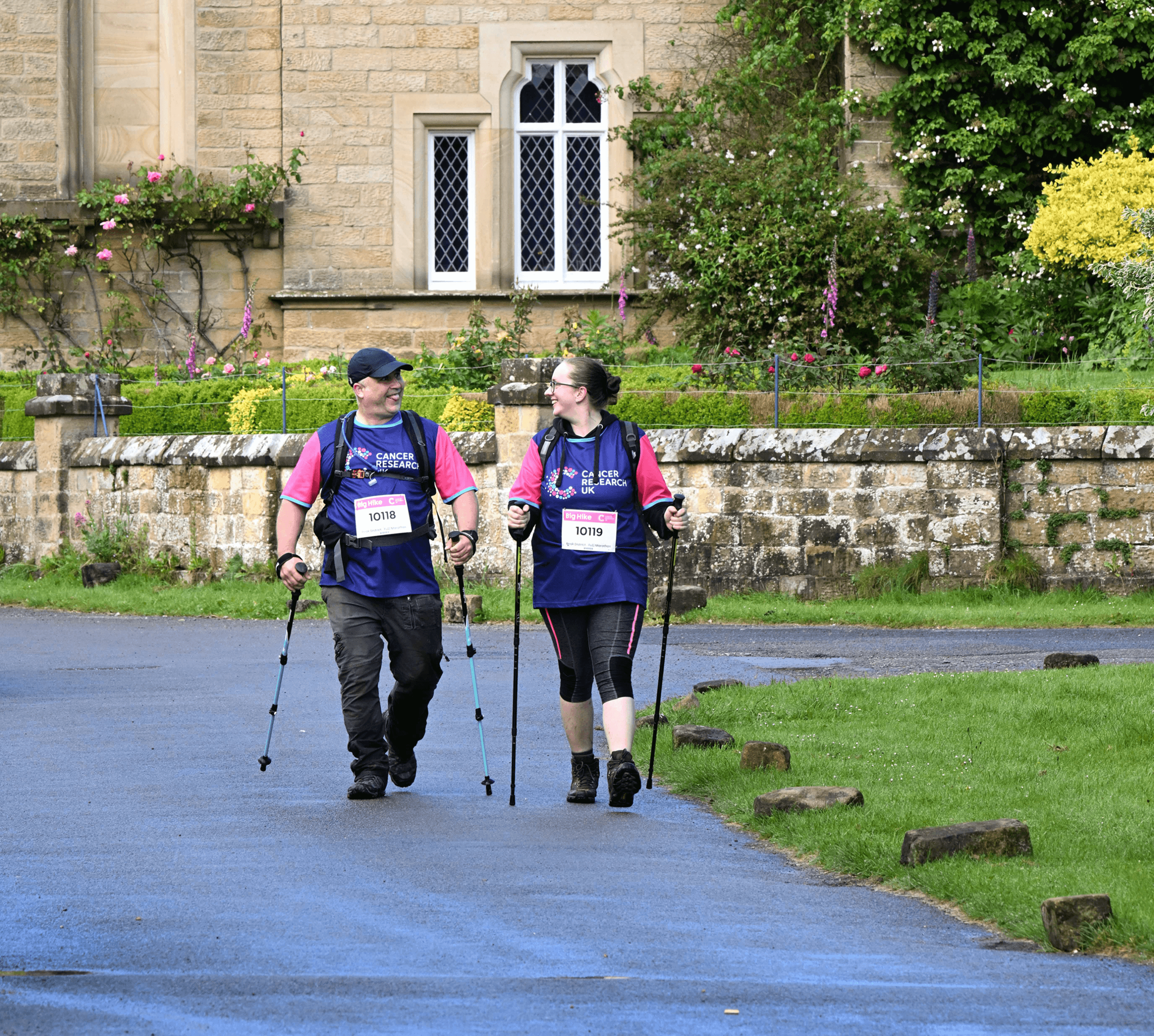 Two Big Hike participants hiking through a village using walking poles wearing Cancer Research UK t-shirts.