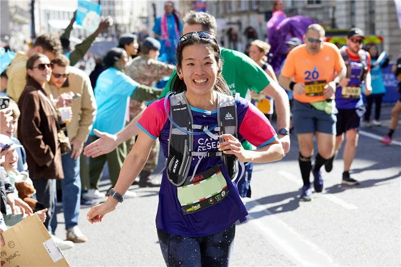 Smiling woman running in a half marathon for Cancer Research UK, with supporters cheering in the background.