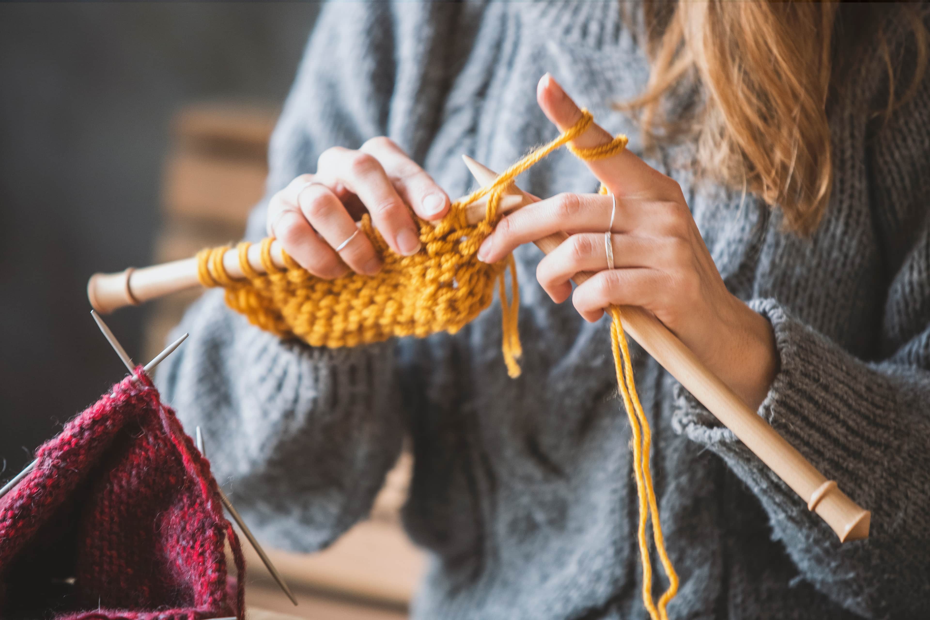 Person knitting with yellow yarn on wooden needles, wearing grey sweater, red knitted piece in foreground.