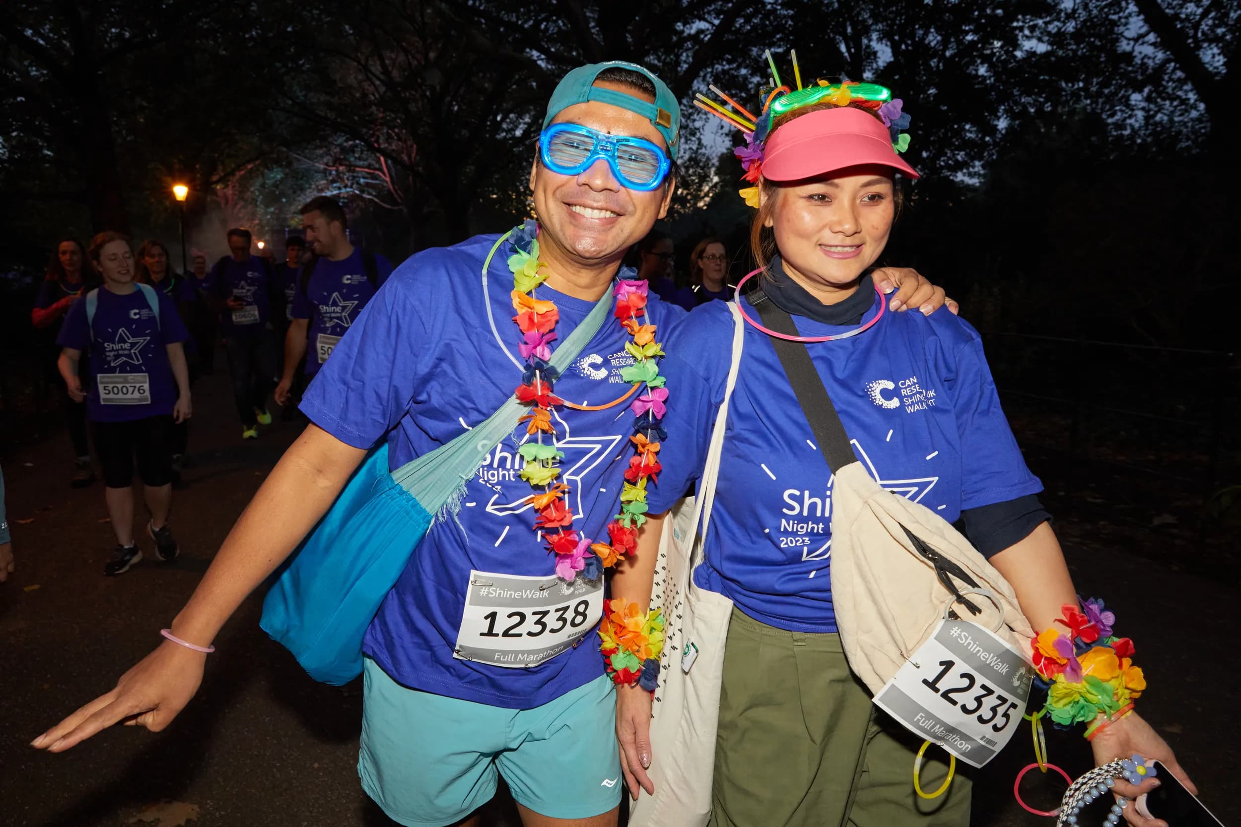 A man and woman wearing Shine Night Walk t-shirts and colourful accessories. They are walking and smiling to the camera.