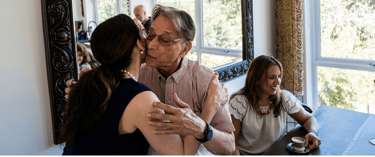 Two people embracing while a third sits nearby at a table It appears to be a family gathering.