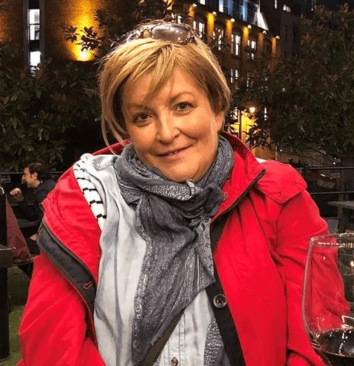Board member Ruth Johnson. Ruth is wearing a red coat and grey scarf, enjoying a drink at an outdoor cafe.