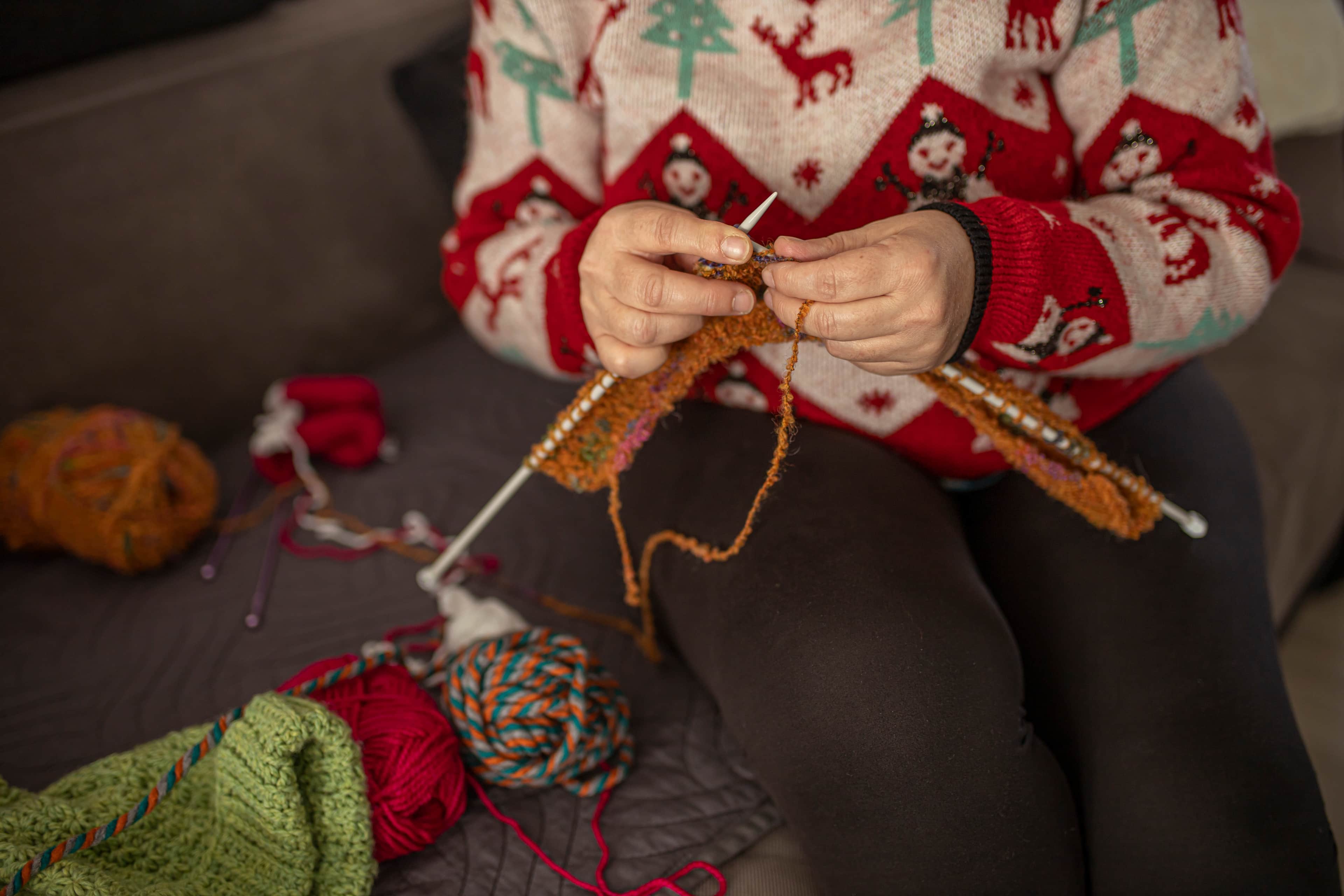 Person knitting with orange yarn on a couch, wearing a festive sweater, surrounded by colourful yarn balls.