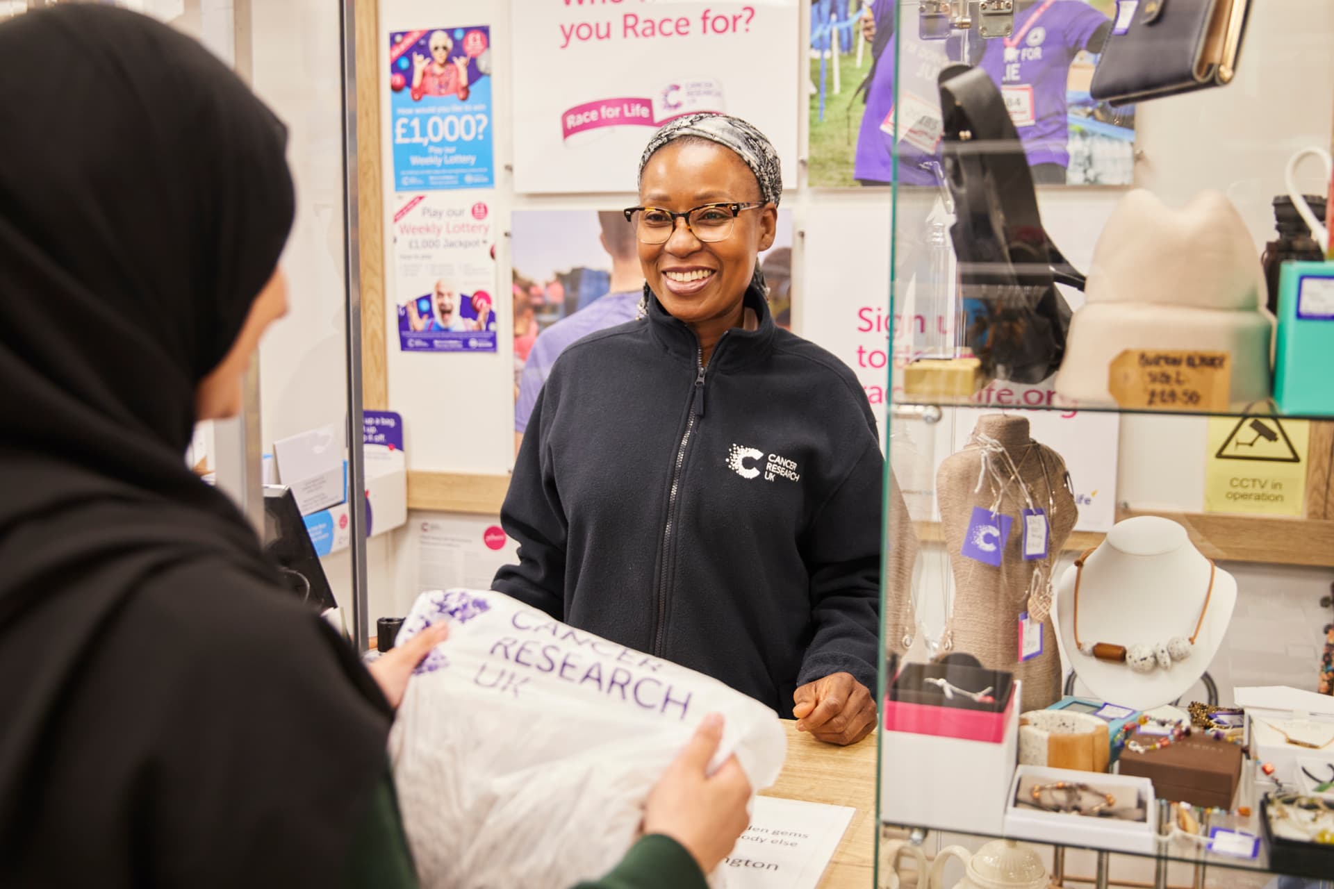 A smiling Cancer Research UK volunteer in a charity shop helping a customer.