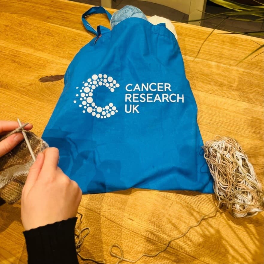 Blue Cancer Research UK tote bag on table with hands knitting beside it.