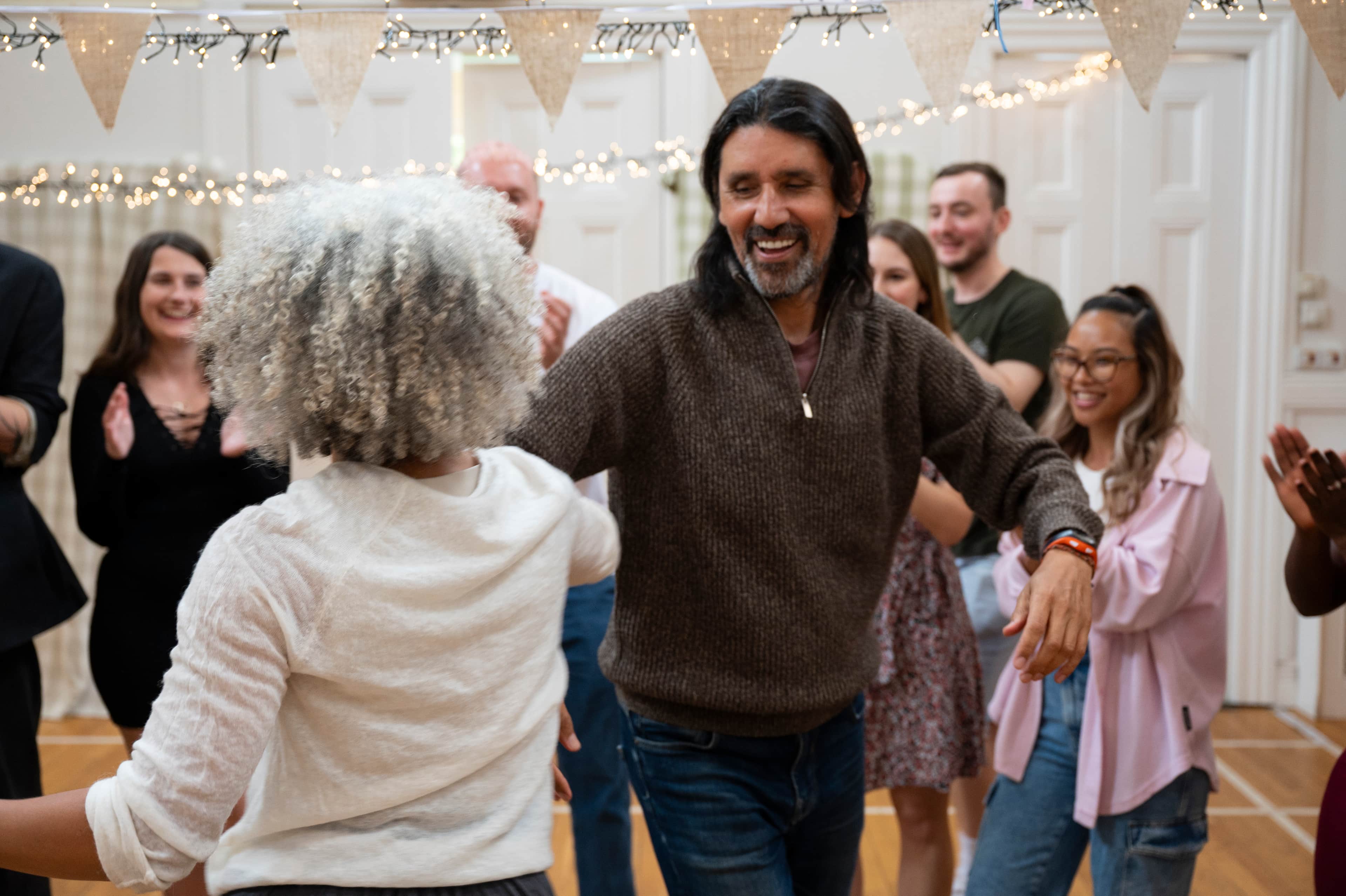 Two people dancing in a decorated hall with bunting and string lights, while others clap in the background.