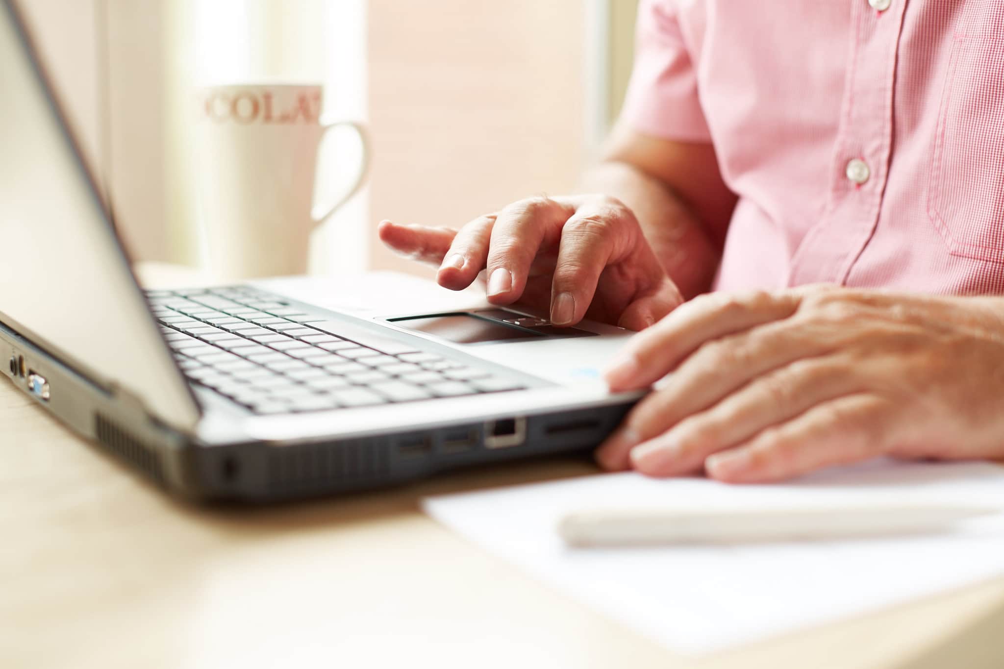 A man is sat at a table, using a laptop. There is a cup of tea besides the laptop.