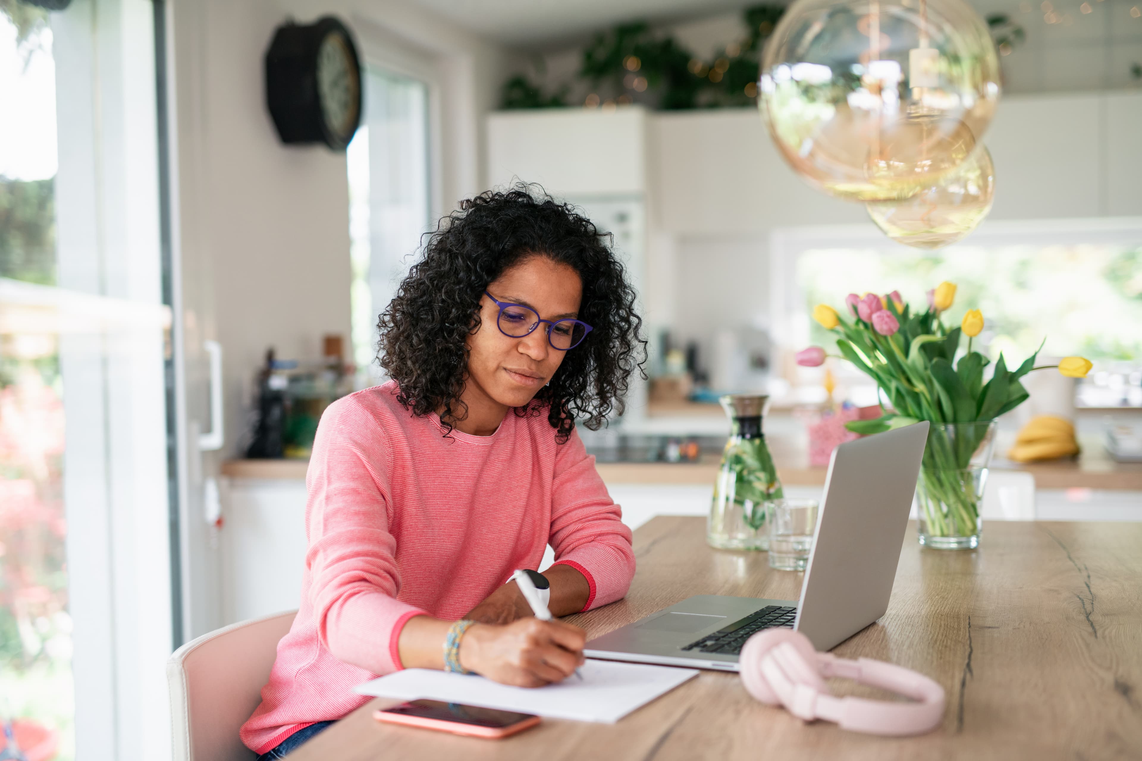 A woman writing information on paper with her laptop in front of her.