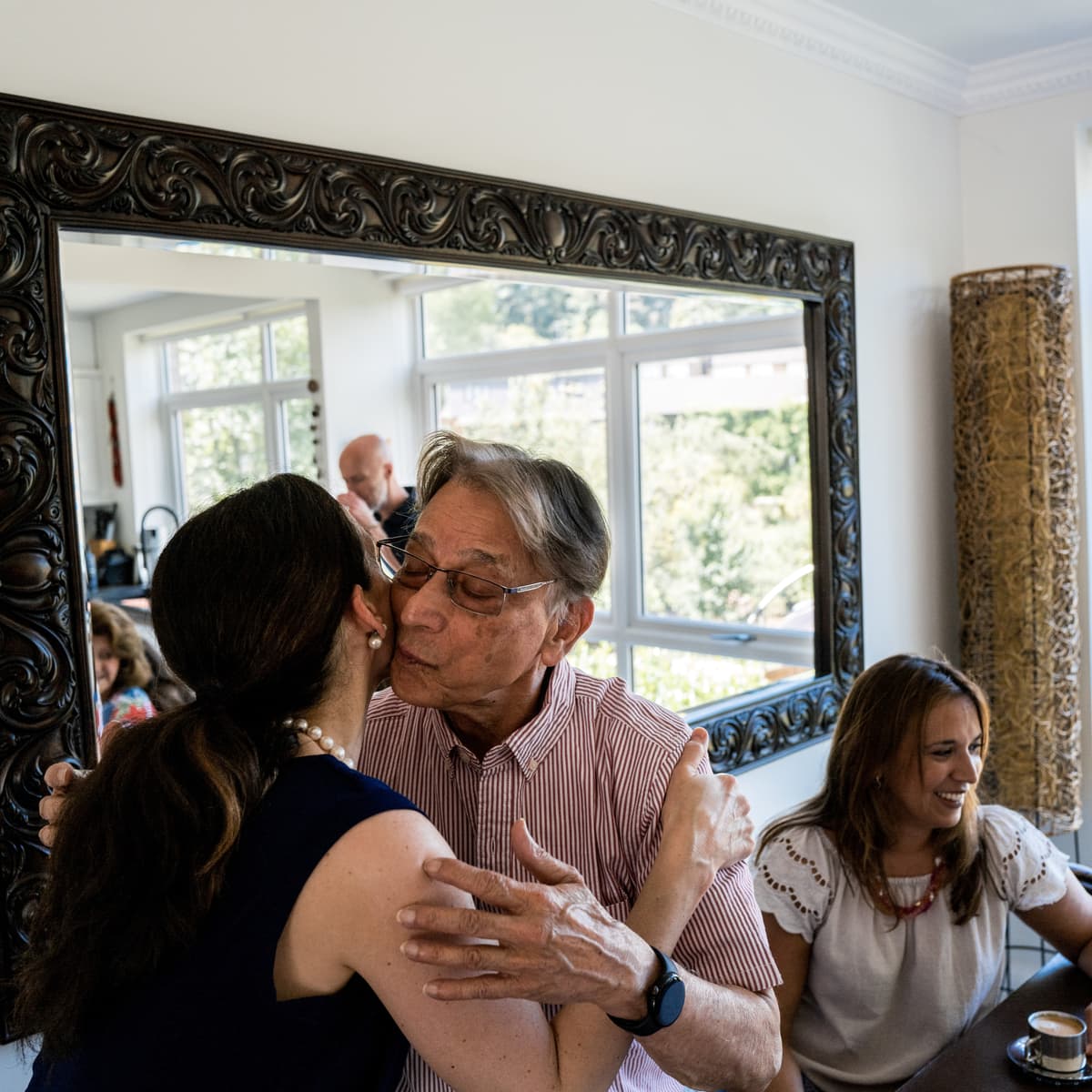 Two people embracing while a third sits nearby at a table. It appears to be a family gathering for lunch.