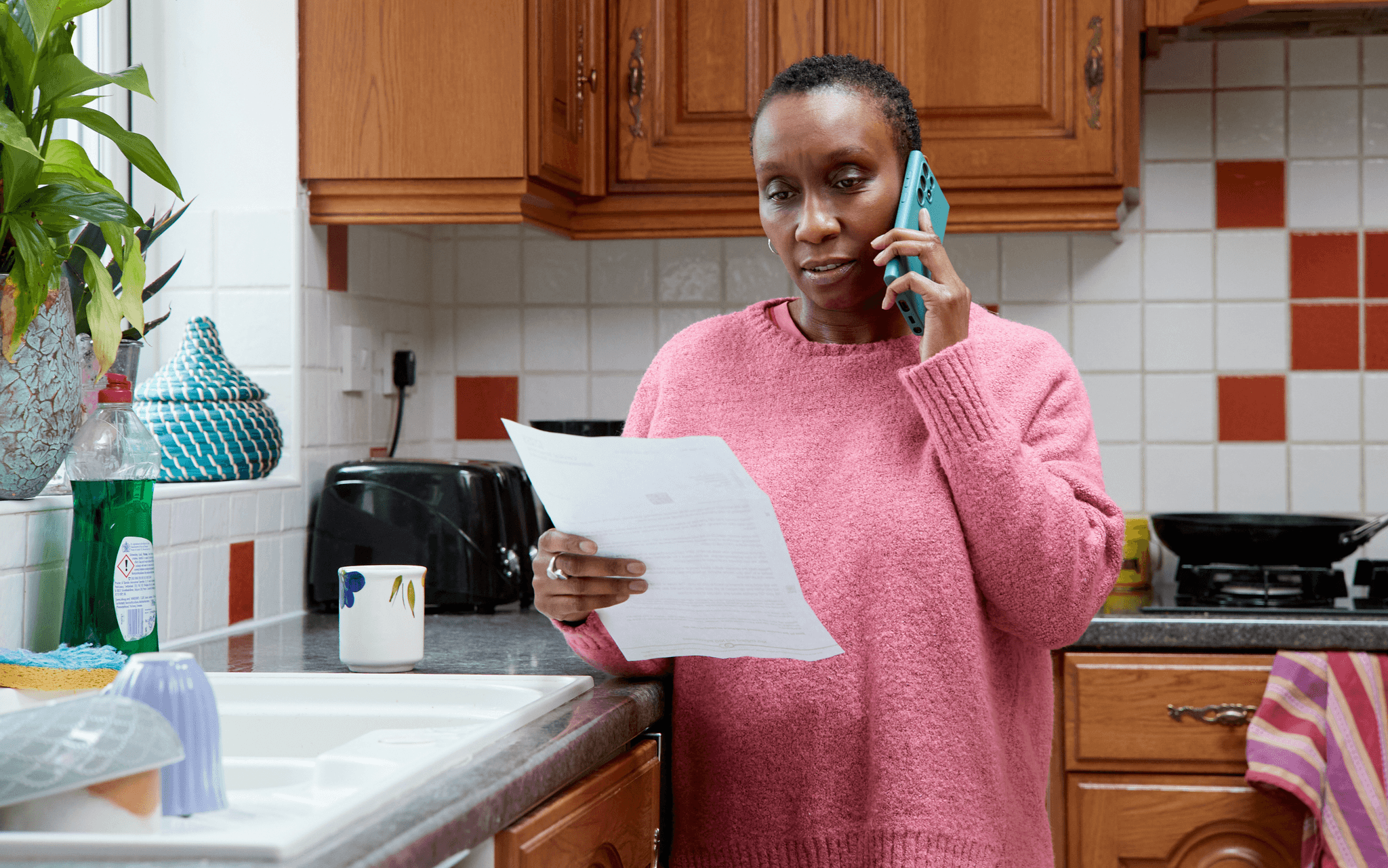 A photo of a woman in the kitchen booking a routine breast or cervical screening appointment on the phone, holding a letter.