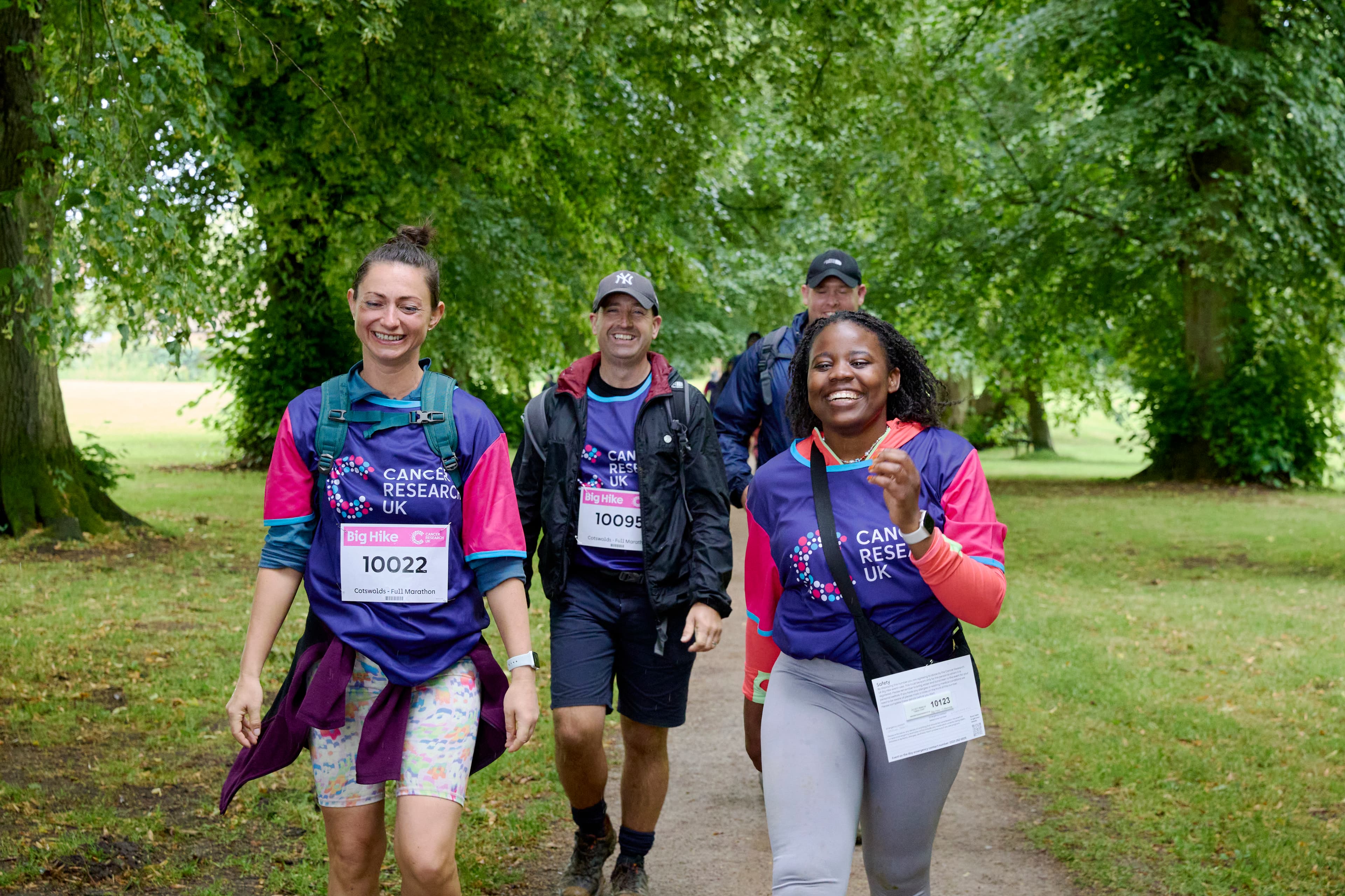 Three Big Hike participants are smilling while walking toward the camera on a tree-lined dirt path.