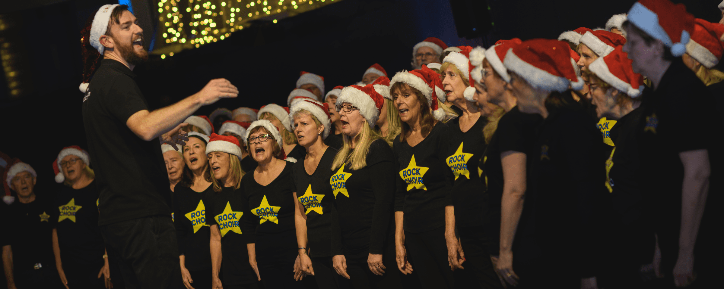 Choir singing with festive hats in front of supporters at a Legacy Festive Music event.