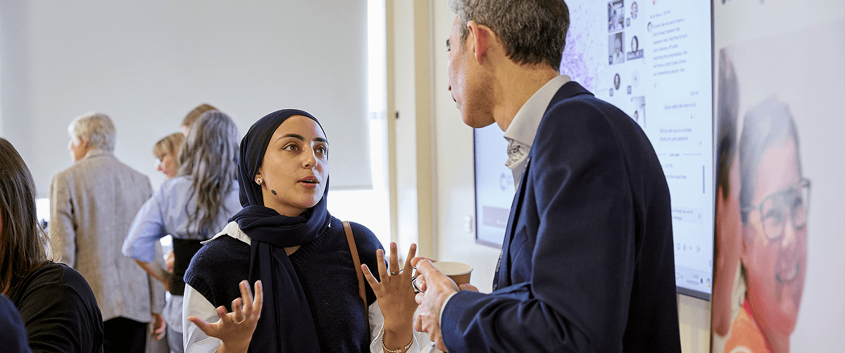 Two people having a discussion at an event. They are standing in front of a screen.