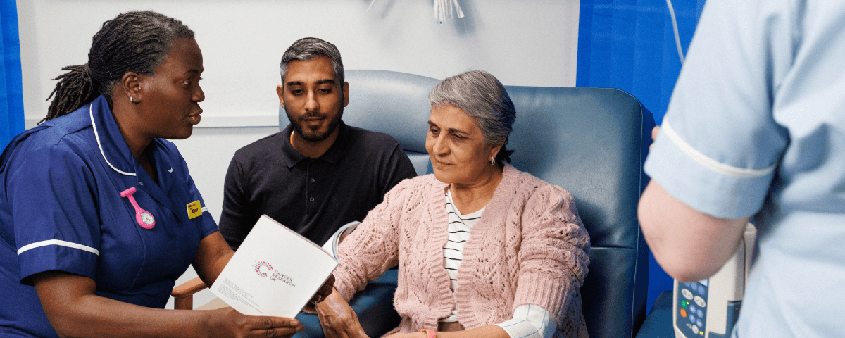 Patient sat with a family member and a health professional while having chemo. Nurse shows them a Cancer Research UK leaflet.