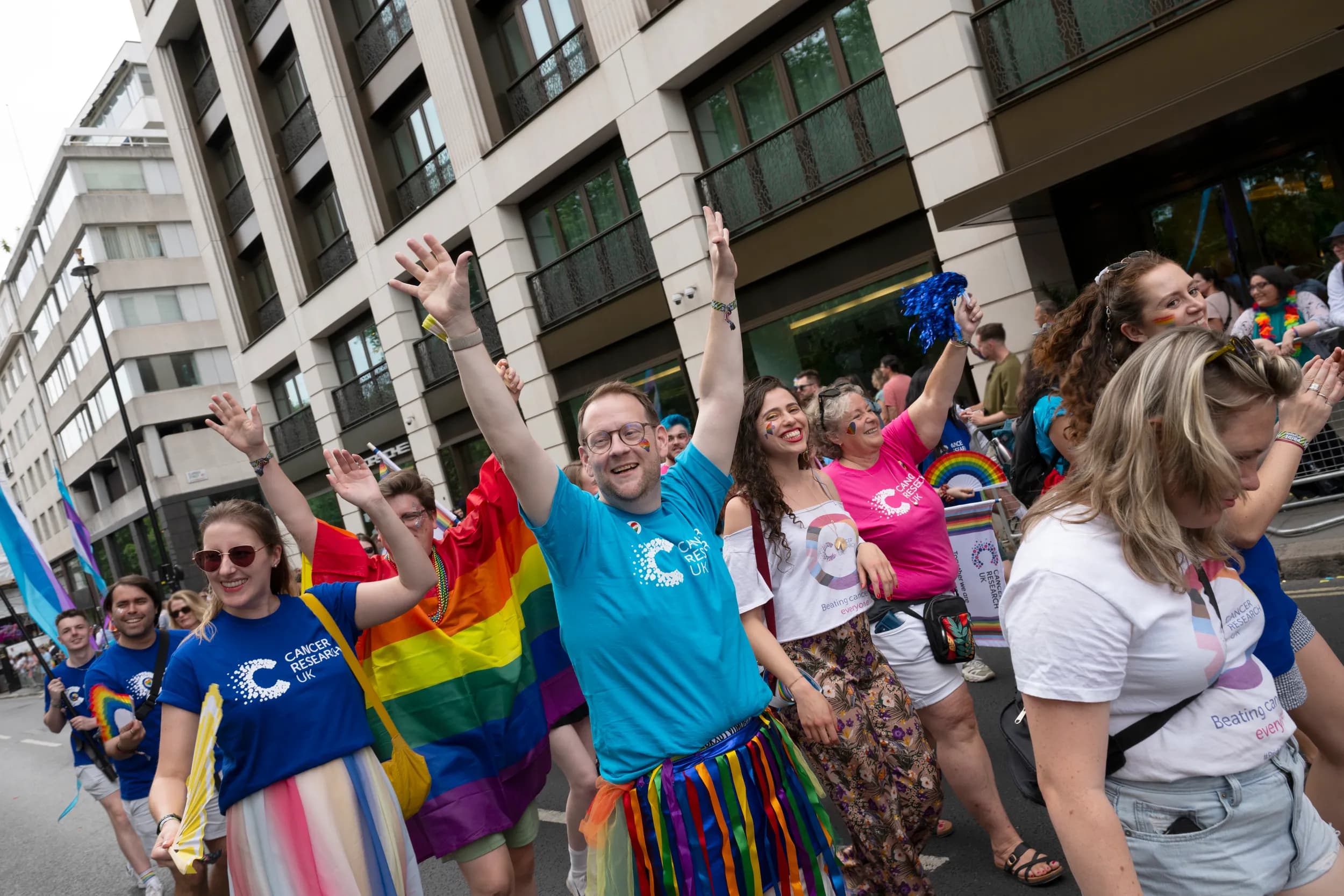 Large group of smiling people cheering and wearing Cancer Research UK t-shirts, walking in a Pride parade.