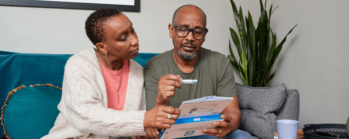 The photo shows a man with his partner on the sofa at home opening a bowel cancer screening test kit from NHS England.