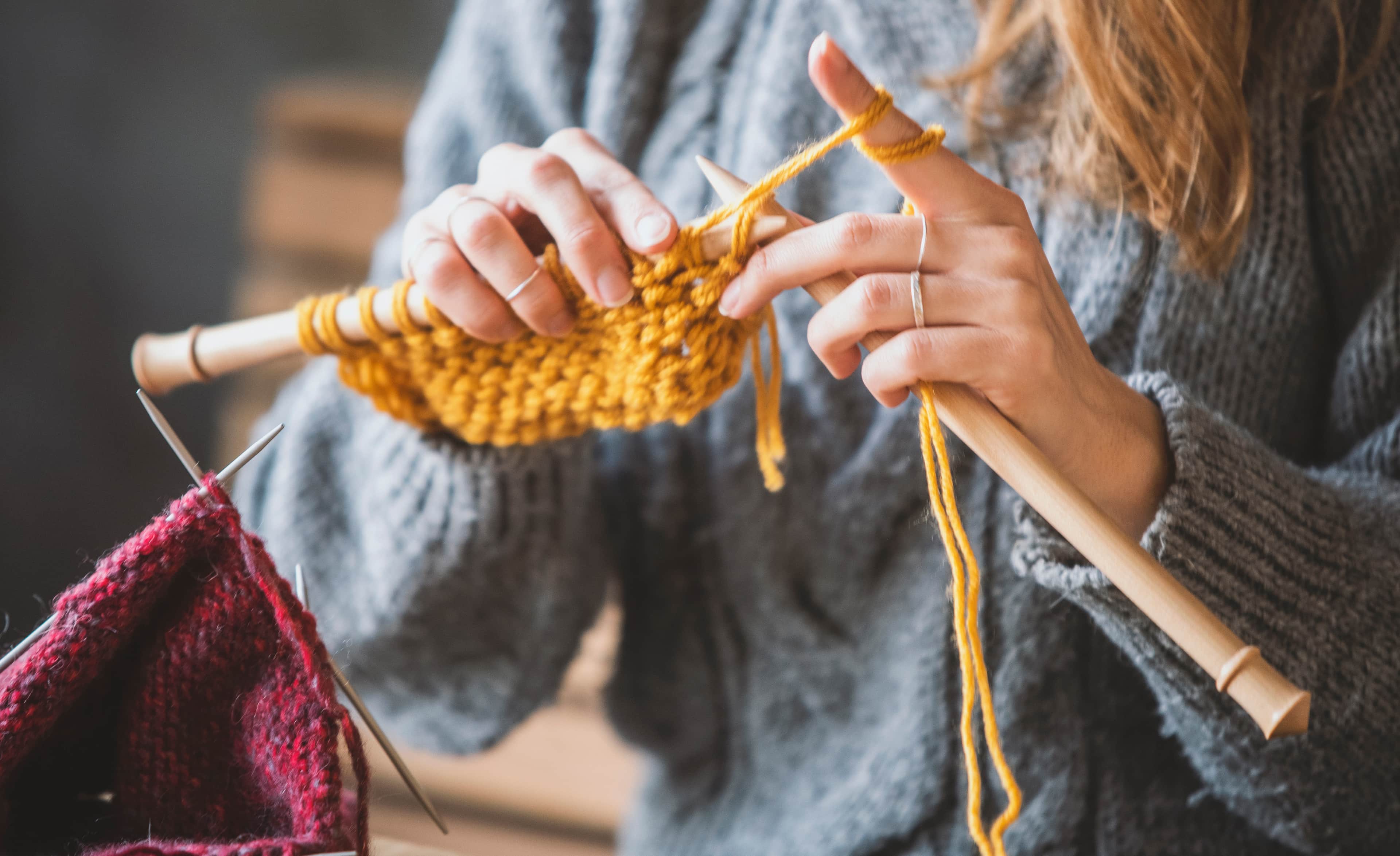 Person knitting with yellow yarn on wooden needles, wearing a grey sweater; red knitting in foreground.