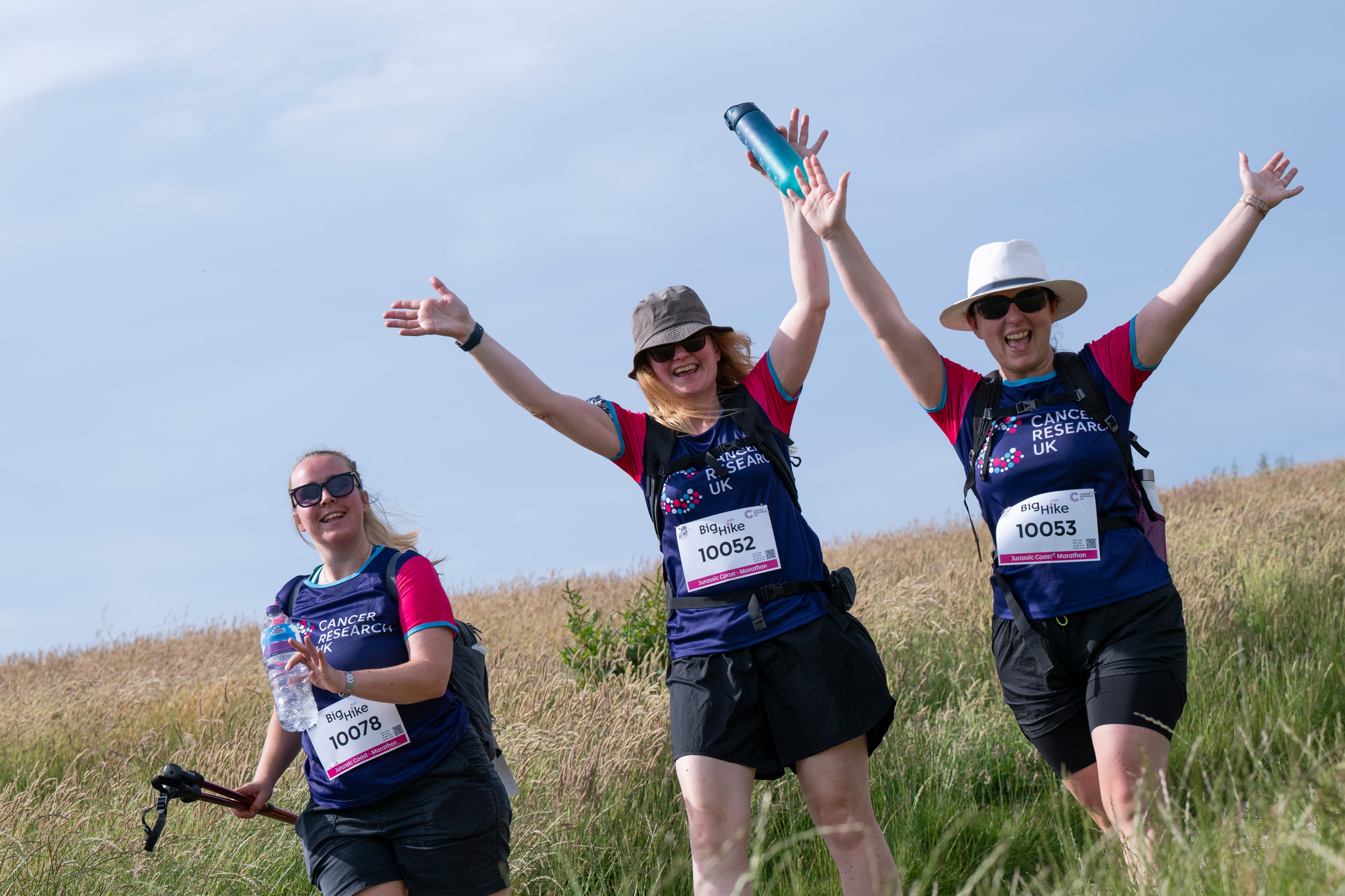Three excited Big Hike participants are cheering with their arms up in the air on a grassy hill.