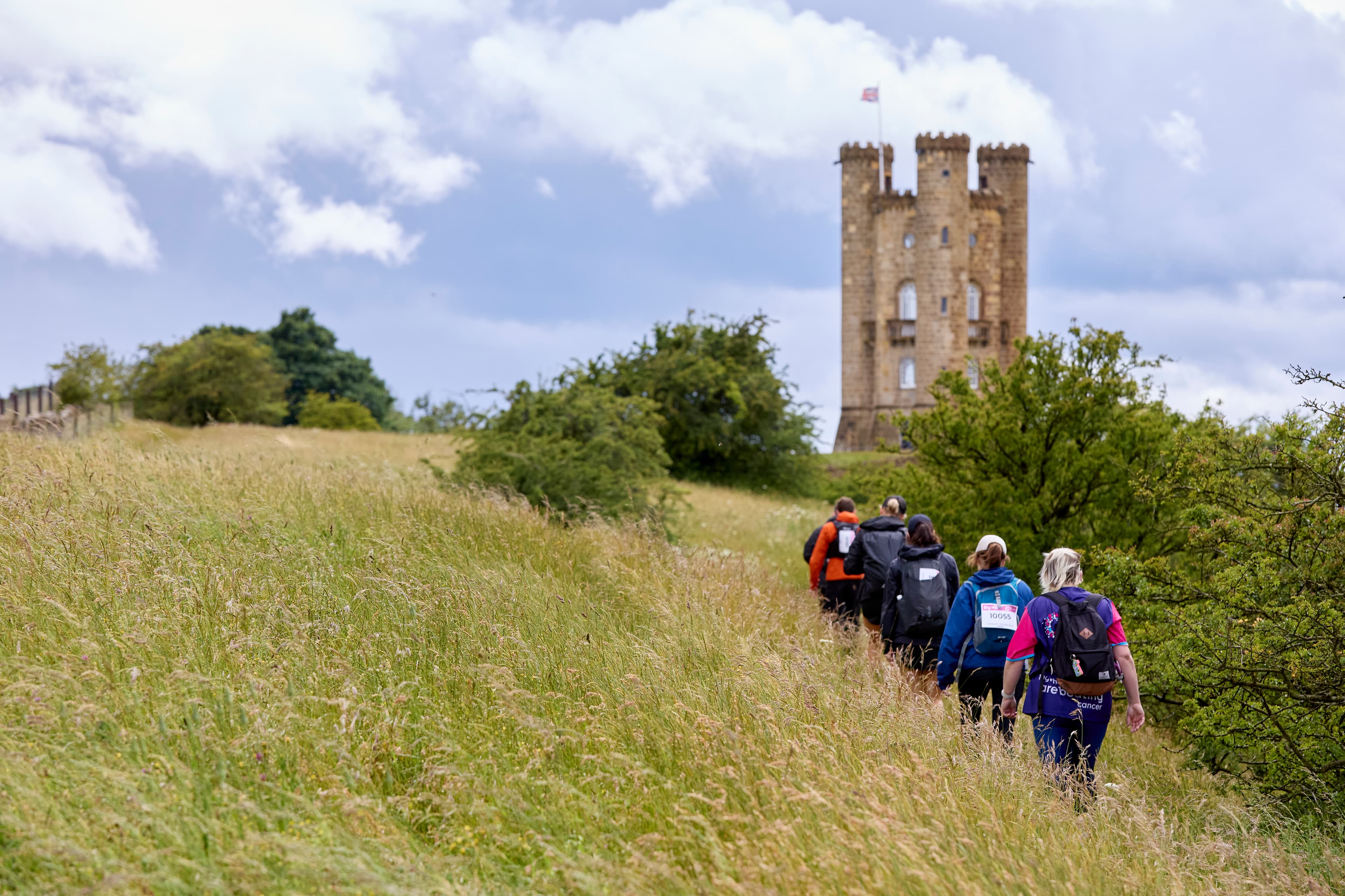 A group of Big Hike participants walk up a grassy hill towards the Broadway Tower under a blue sky.
