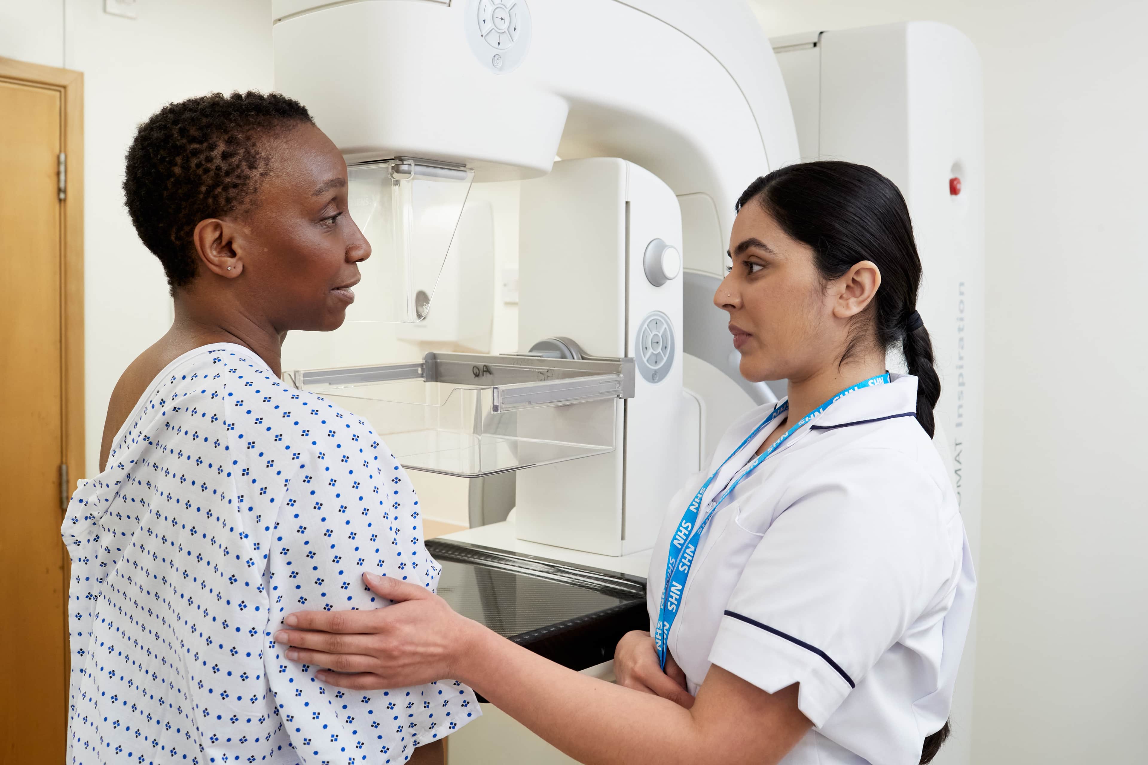 A mammographer speaking to a patient, wearing a hospital gown, before a mammogram.