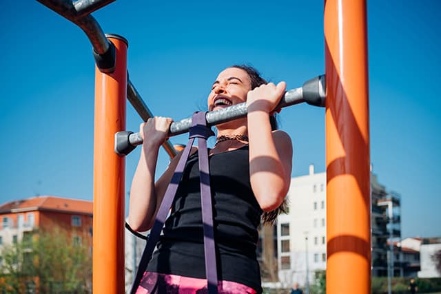 A photo of a smiling woman completing a band-assisted pull-up outdoors using a pull-up station.