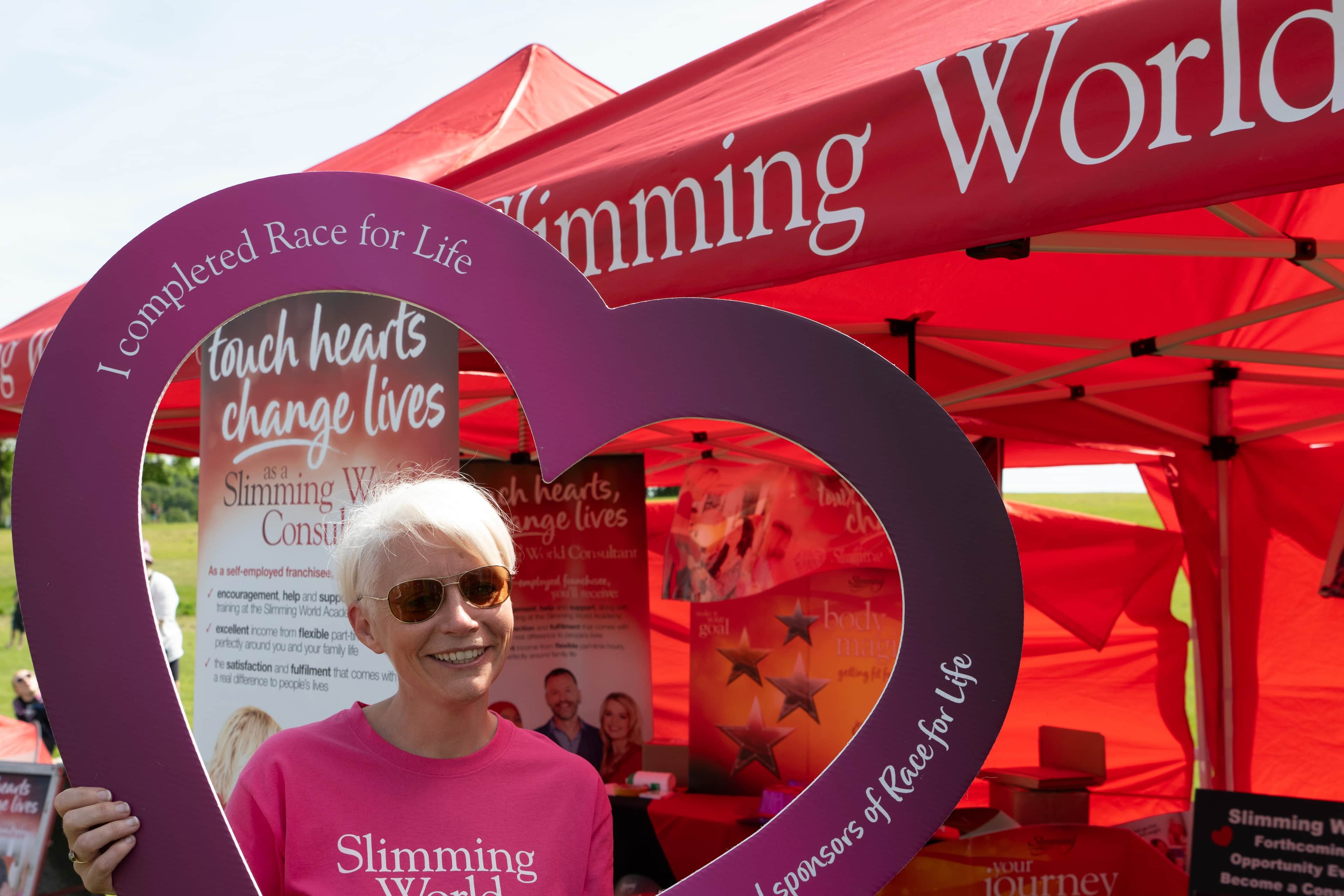 A woman standing outside a 'Slimming World' booth at a Race for Life event. She is smiling and posing with a heart cut-out.