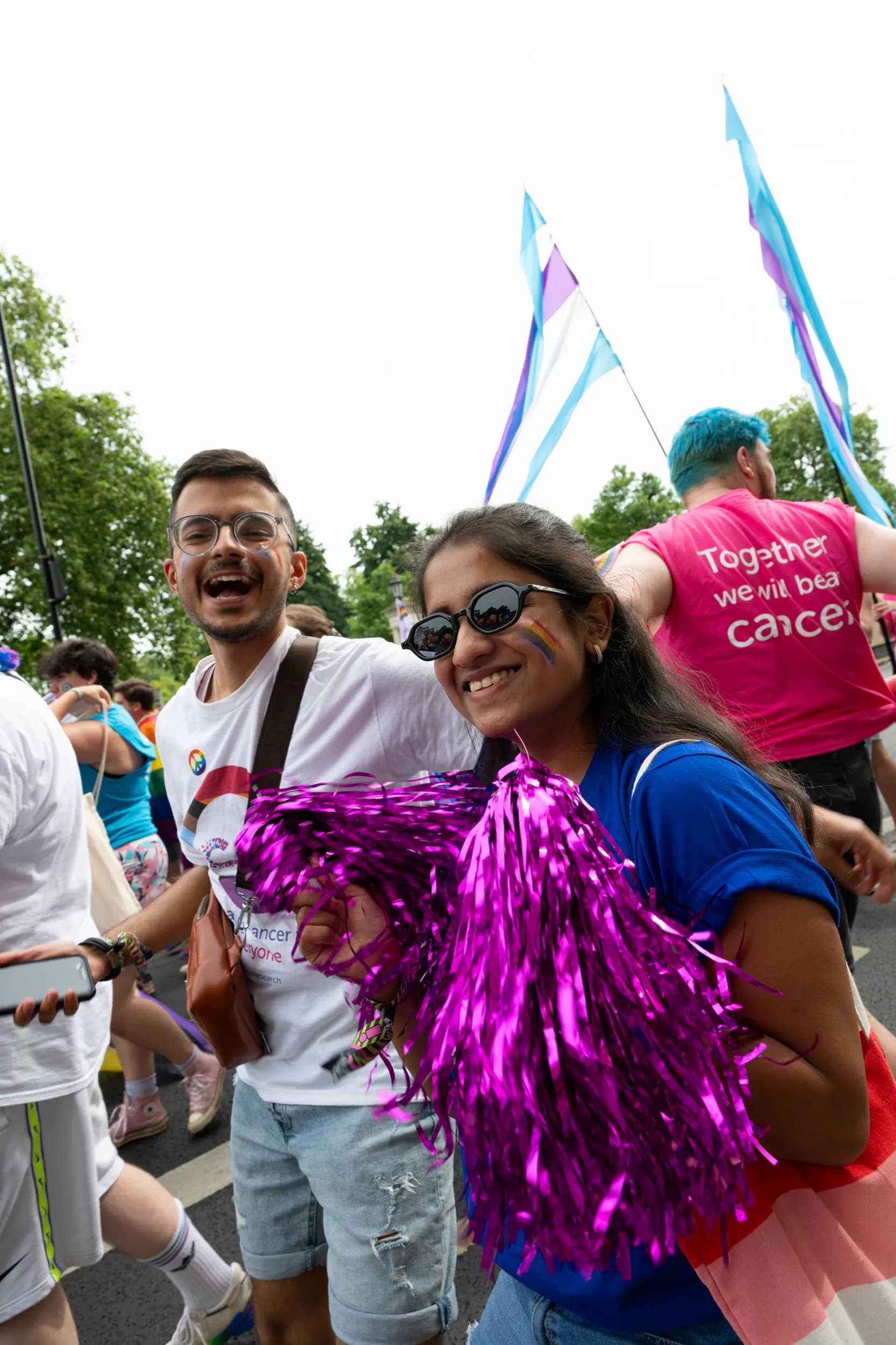 Two people at a Pride parade, smiling and carrying pink pom poms.