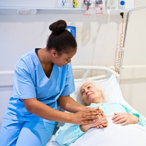 An older woman is lying in a hospital bed, with a healthcare professional sat next to her comforting her.
